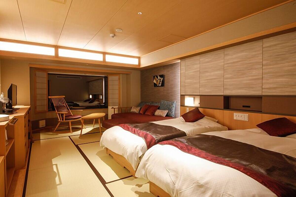 Habitación de hotel con suelo de tatami, una cama de estilo japonés tradicional, un escritorio de madera y una puerta corredera que da a un balcón.