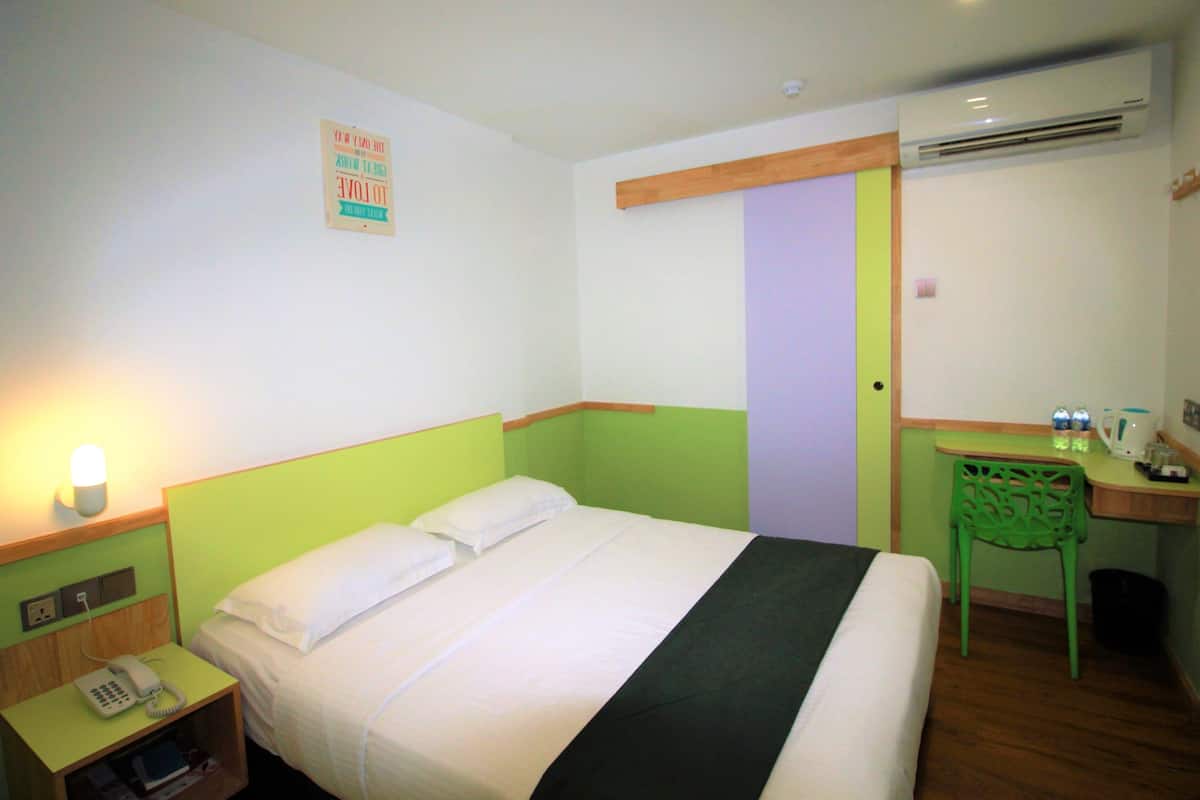 A hotel room with a bed, bedside table with a phone, a green chair, a desk with a kettle, and an air conditioning unit.