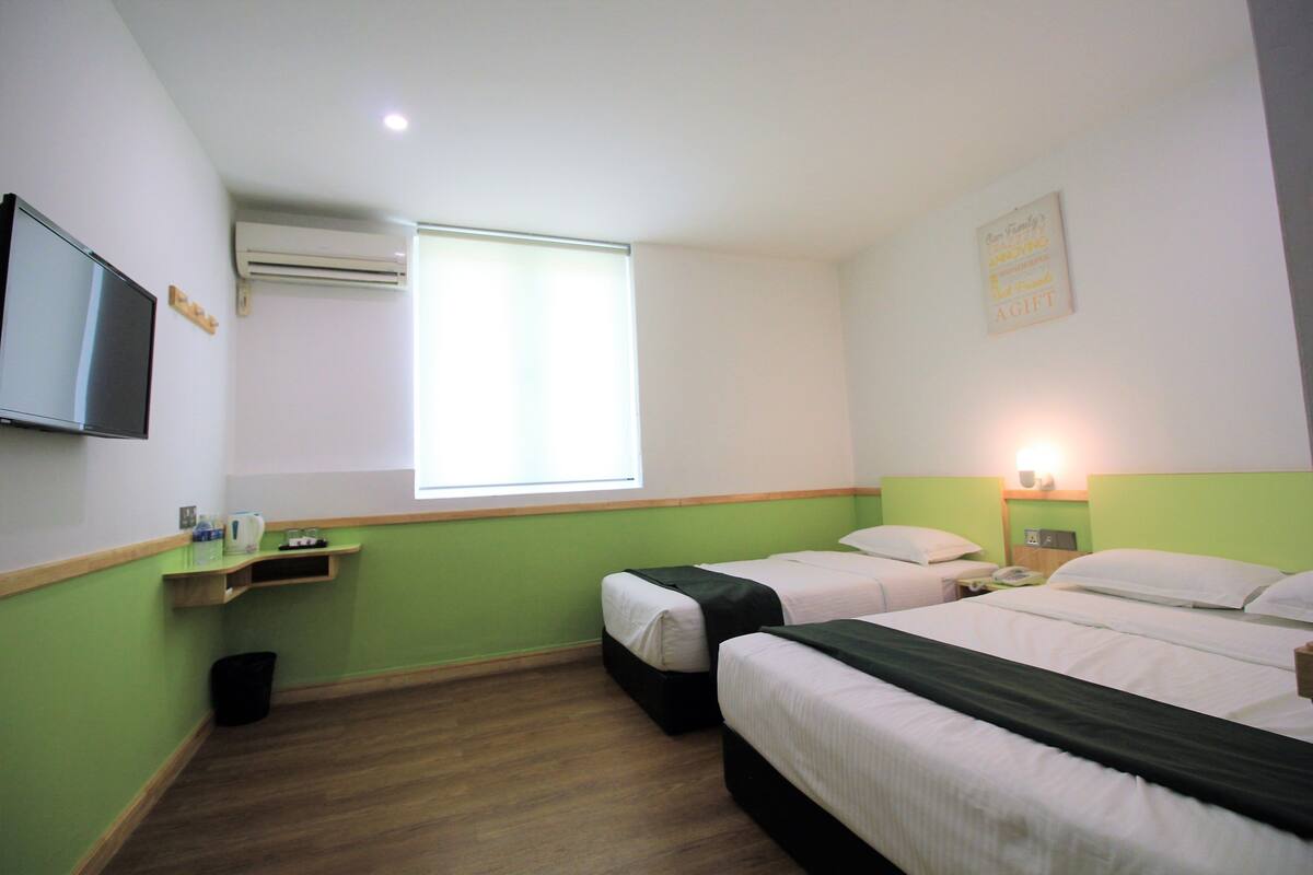A hotel room with two beds, a TV, an air conditioner, a window, and a bedside table with a lamp.