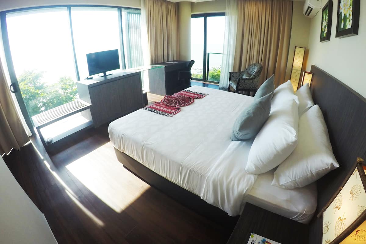 A hotel room with a large bed, a desk with a TV, a chair, and a balcony with a view of greenery.
