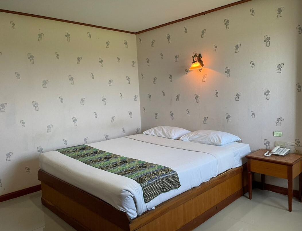 Una cama individual con estructura de madera, ropa de cama blanca y una manta estampada. Se ve una mesita de noche con una lámpara y un teléfono.