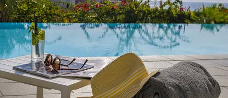 A poolside setting with a table, a rolled towel, a straw hat, and a drink with a straw and mint garnish.