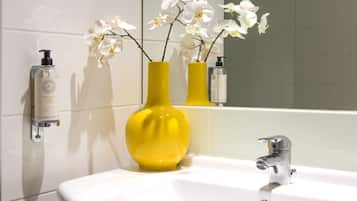 A bathroom with a white sink, a yellow vase with white flowers, and a mirror reflecting the sink and vase.