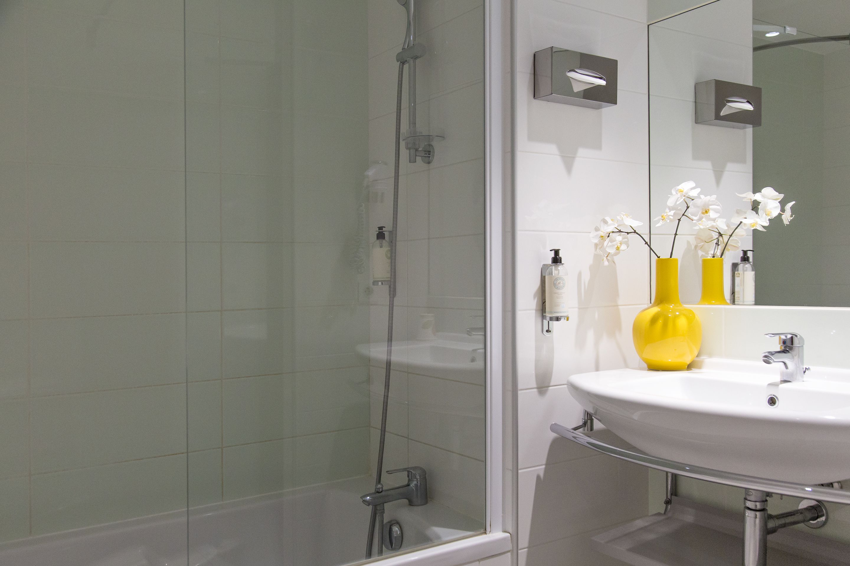 A bathroom with a glass-enclosed shower, a white sink, and yellow vases with white flowers.