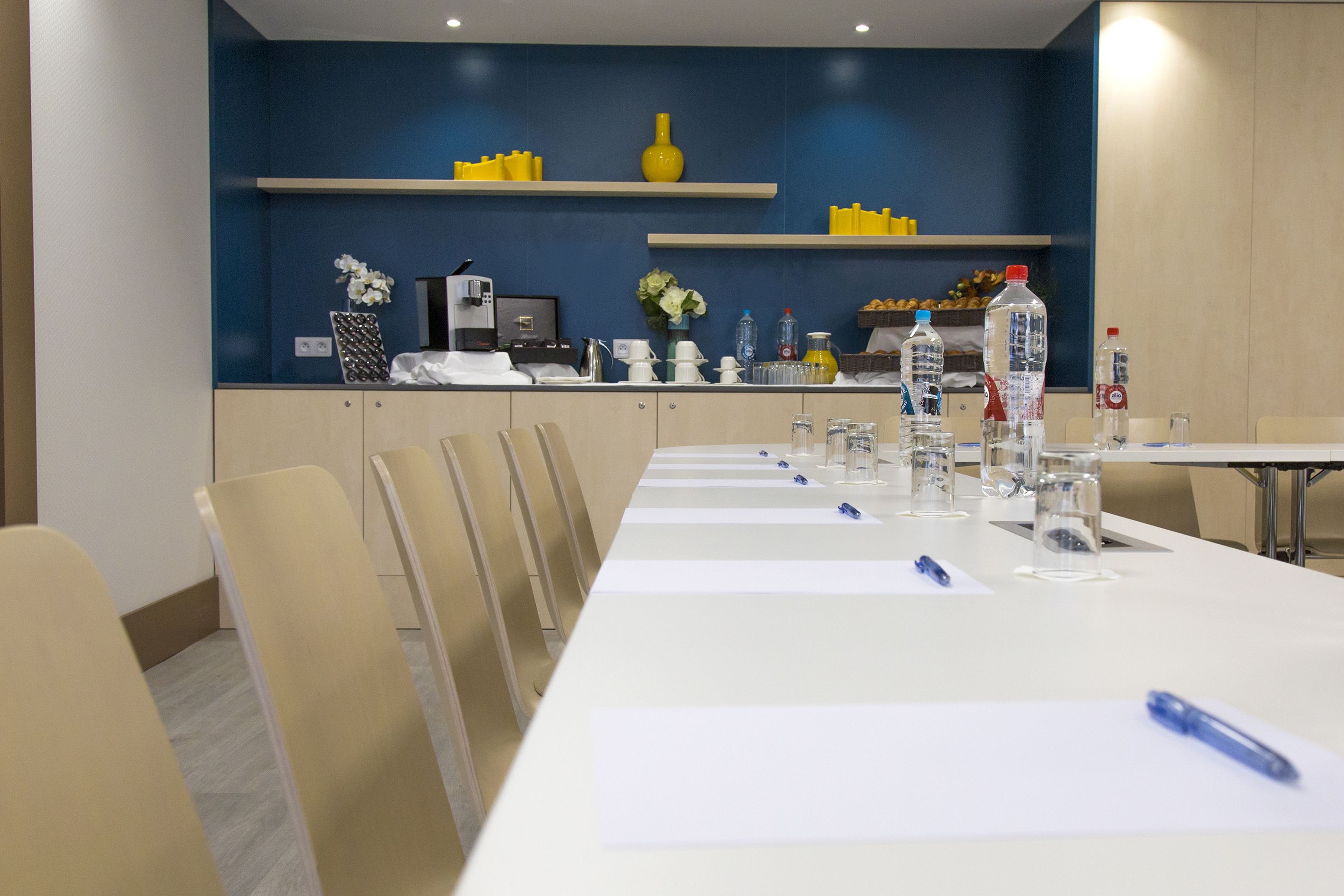 Un salón de conferencias con una larga mesa, sillas y varios servicios como botellas de agua, vasos y una máquina de café.