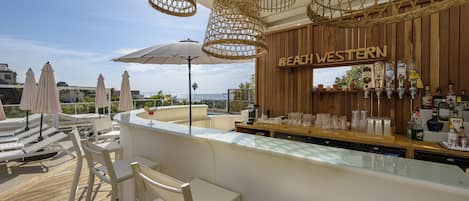 A beach-themed bar area with white bar stools, wicker hanging lights, and a view of the sea.
