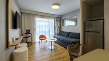 Ett modernt hotellrum med en soffa, ett litet runt bord, en tv som är monterad på väggen, ett kylskåp och ett trägolv.