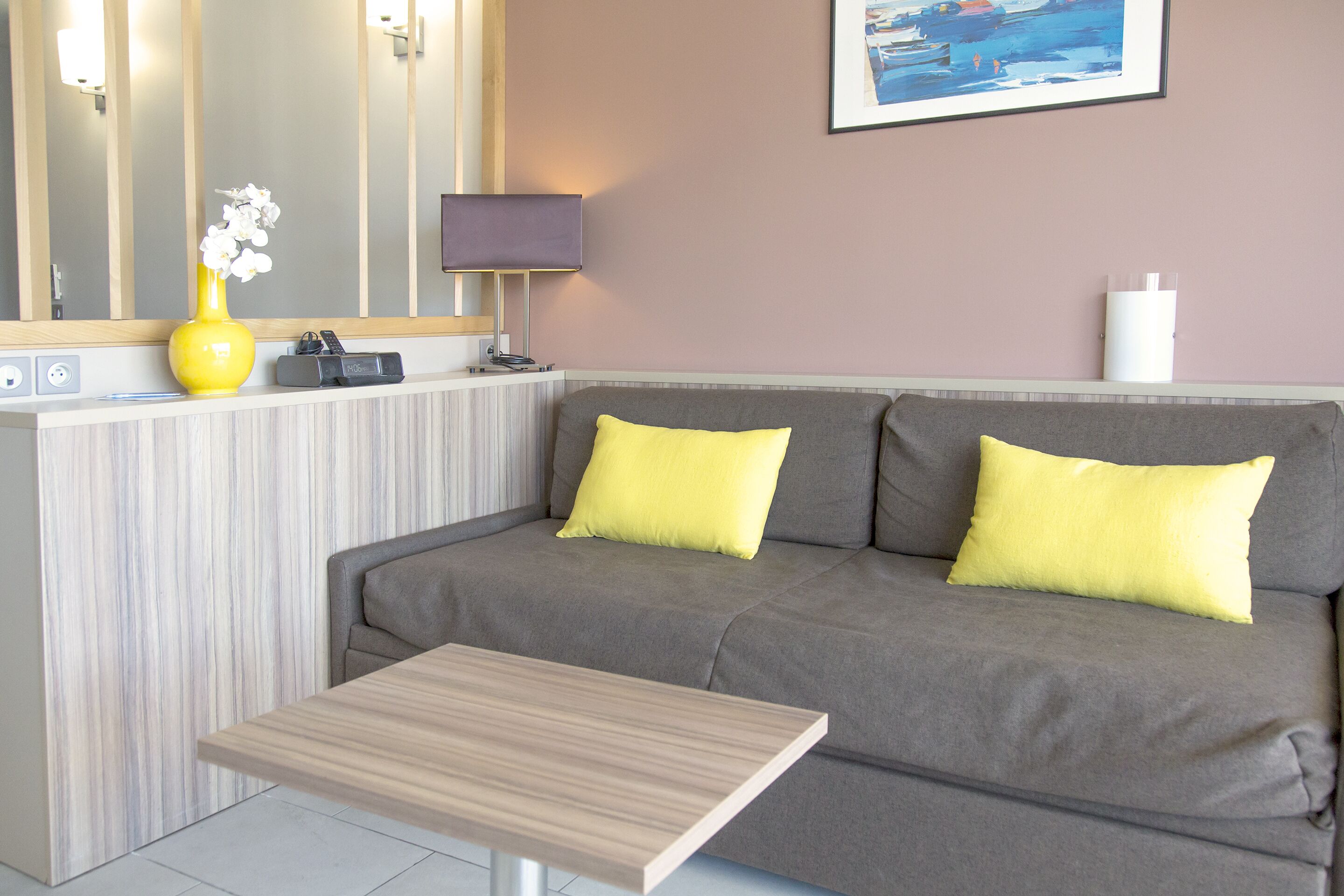 A modern living room with a grey sofa, two yellow cushions, a wooden coffee table, a yellow vase with white flowers, a lamp, and a framed painting on the wall.