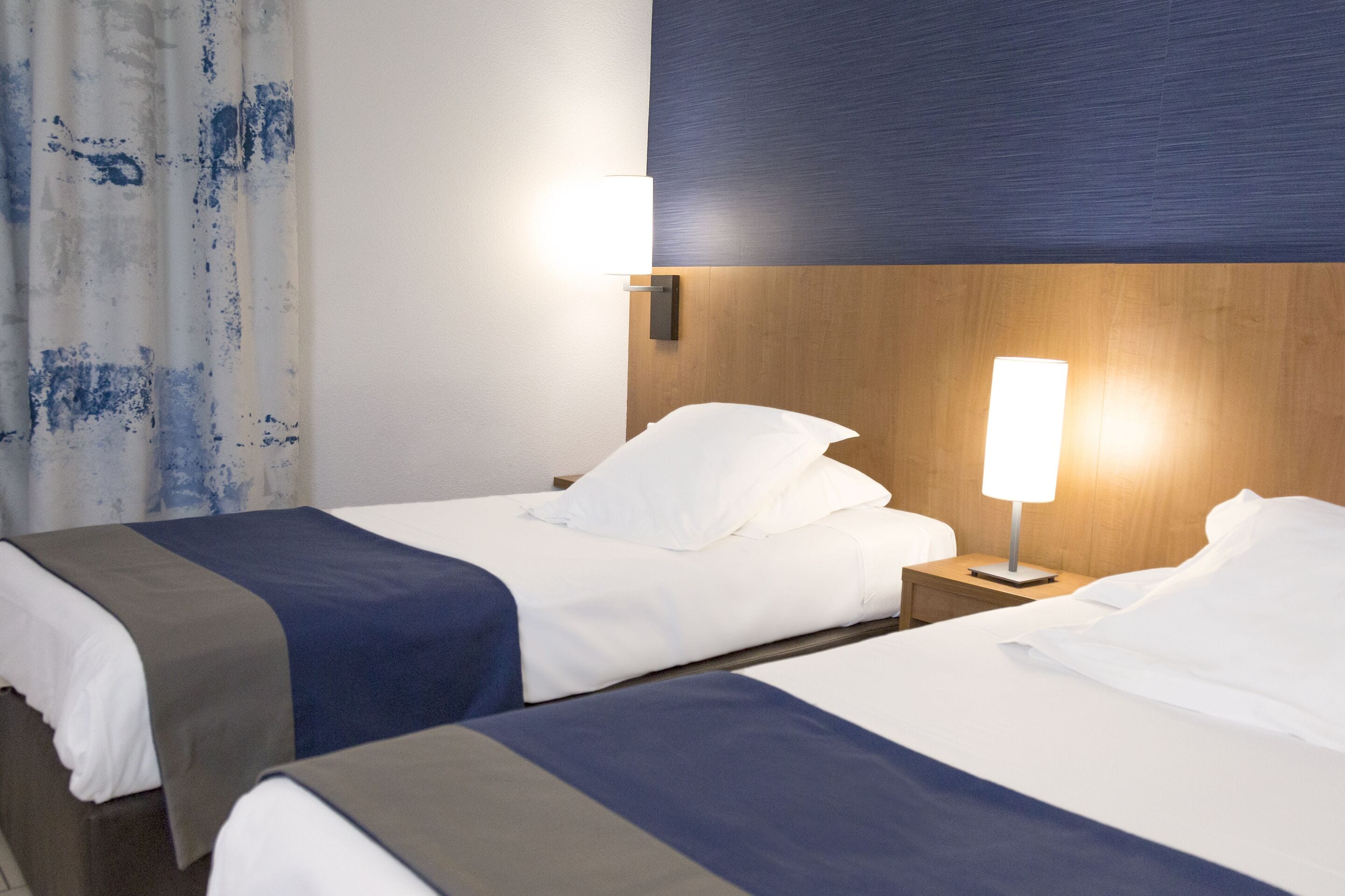 Camera d'albergo con due letti, entrambi con lenzuola bianche e un tocco di blu. Sono presenti comodini con lampade e una testiera in legno.
