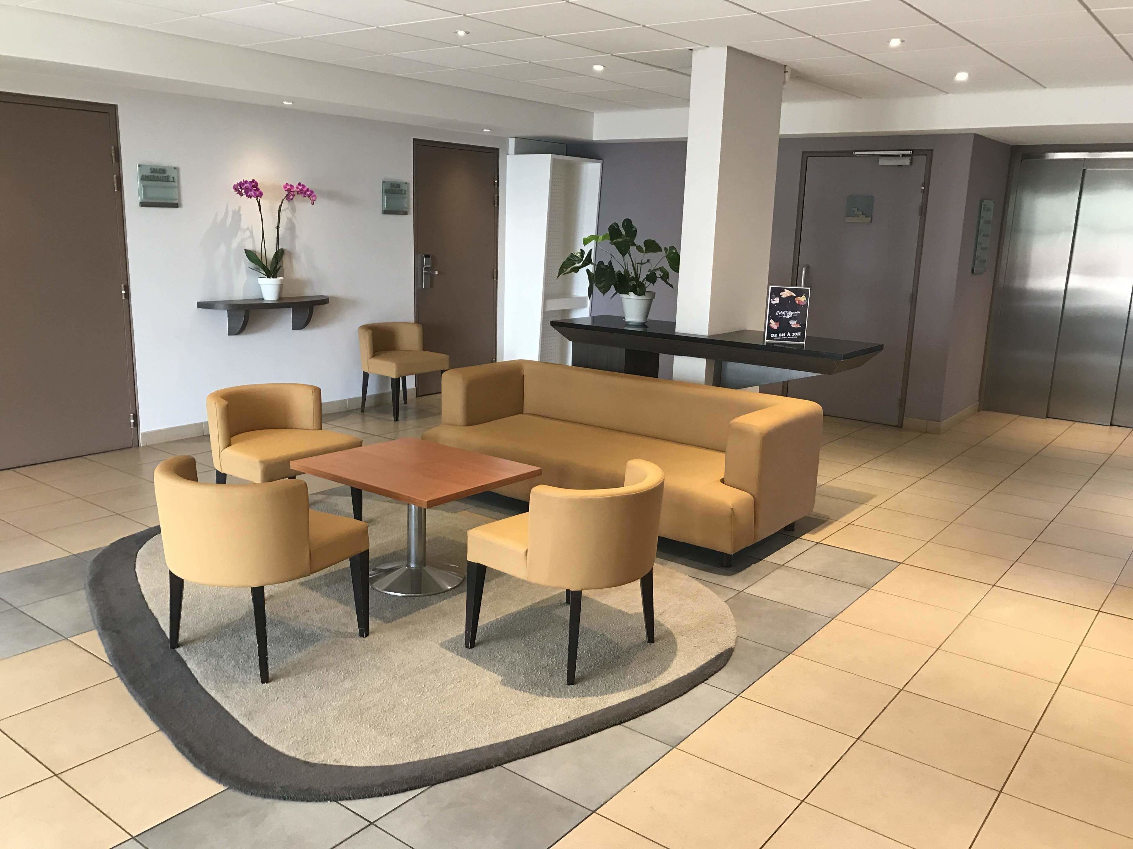 A lobby with a beige sofa, armchairs, a coffee table, a potted plant, a small table with a cup, and a sign on the wall.