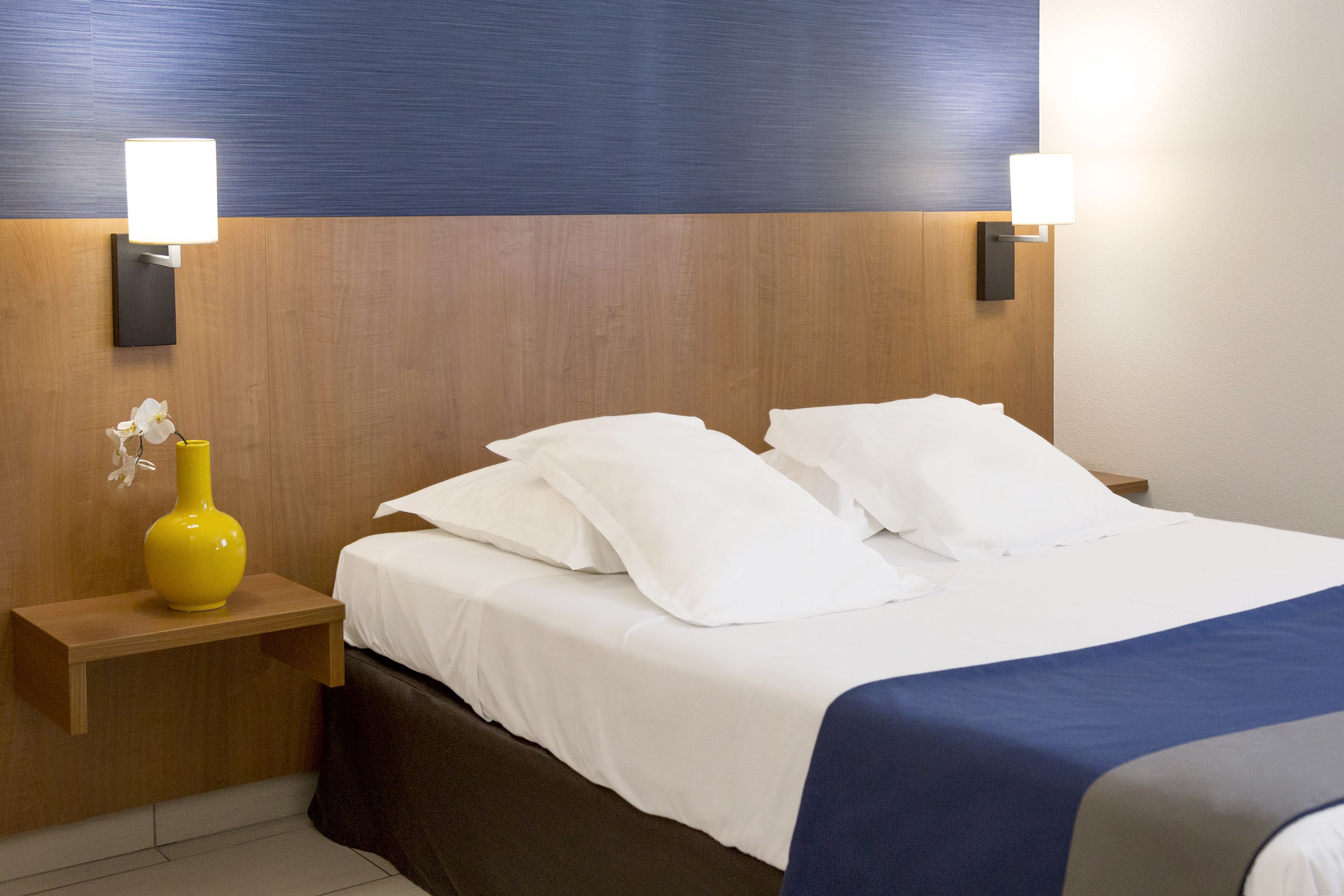 Hotellihuone, jossa on puinen sängynpääty, valkoisilla lakanoiden peitetty sänky, keltainen maljakko yöpöydällä ja seinään kiinnitetyt valaisimet.