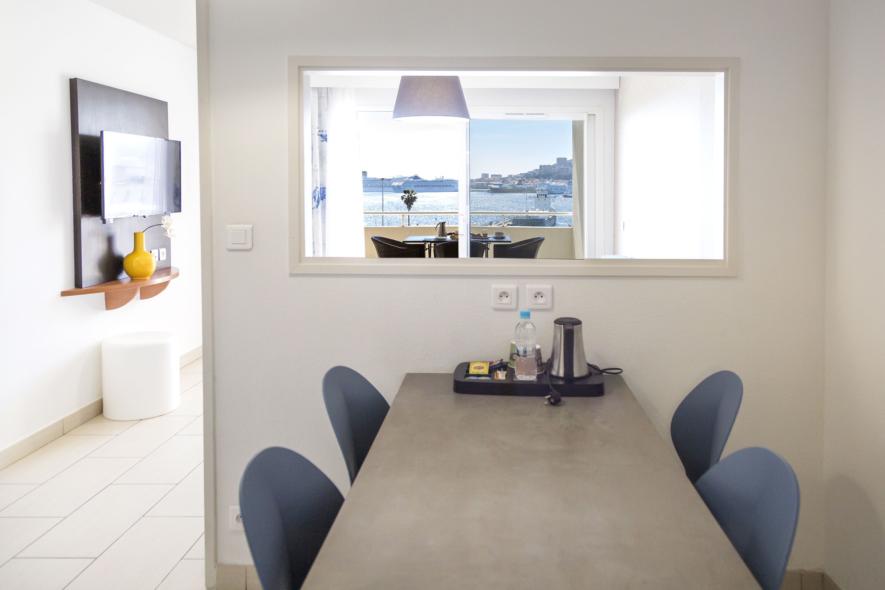 Un comedor con vistas al mar, una mesa con sillas, una lámpara y una pequeña estantería con un jarrón y una planta.