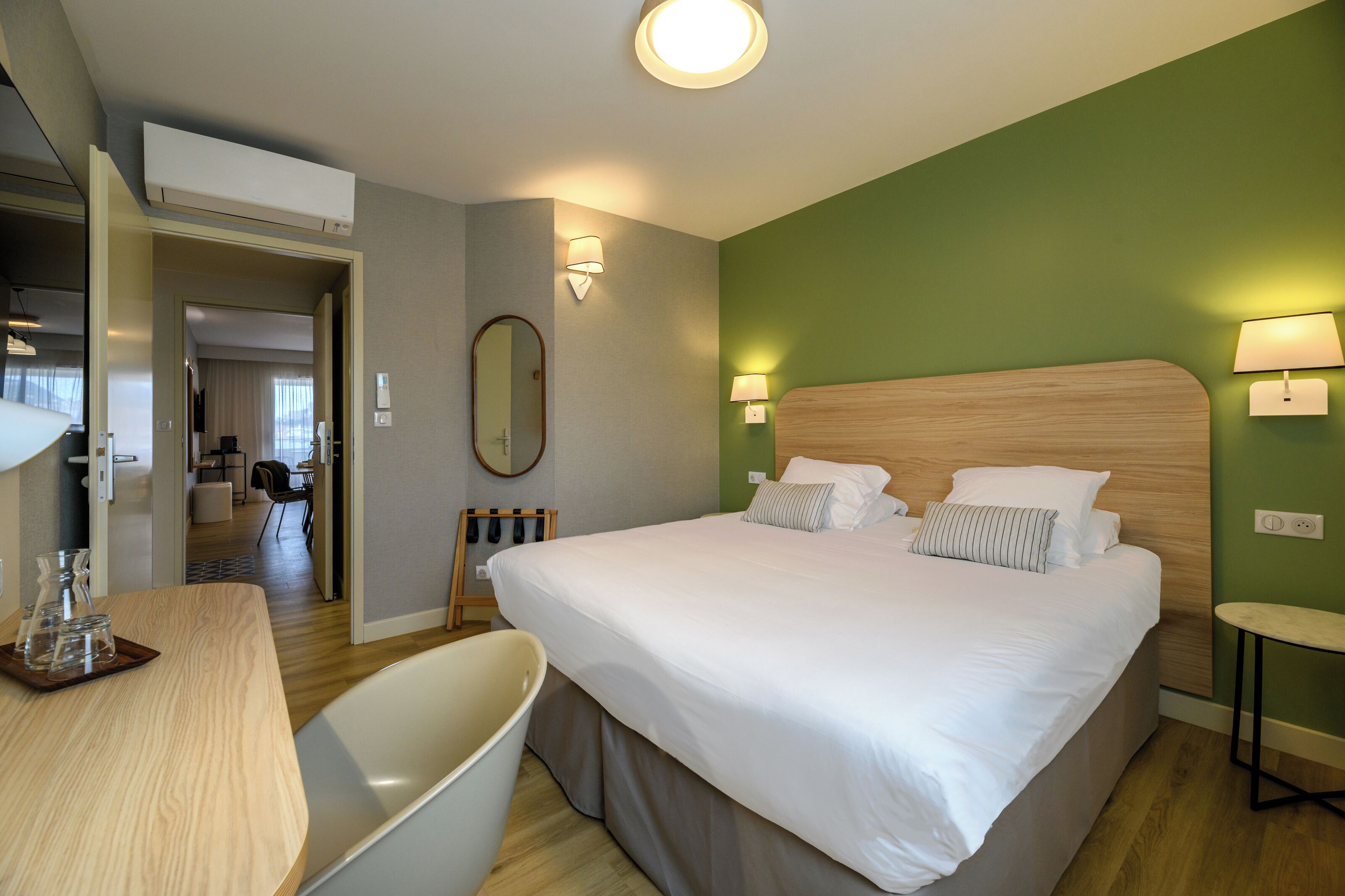 A hotel room with a large bed, bedside lamps, a wooden headboard, a desk with a chair, and a mirror on the wall.