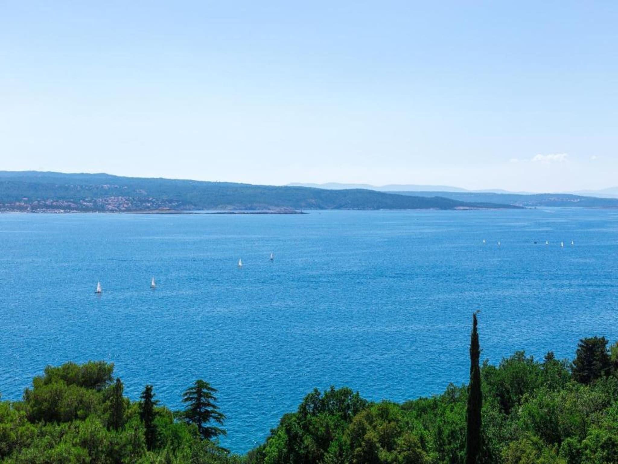 Une vue sur la côte, avec une mer bleue et limpide, plusieurs voiliers et une ligne de côte lointaine parsemée de bâtiments.