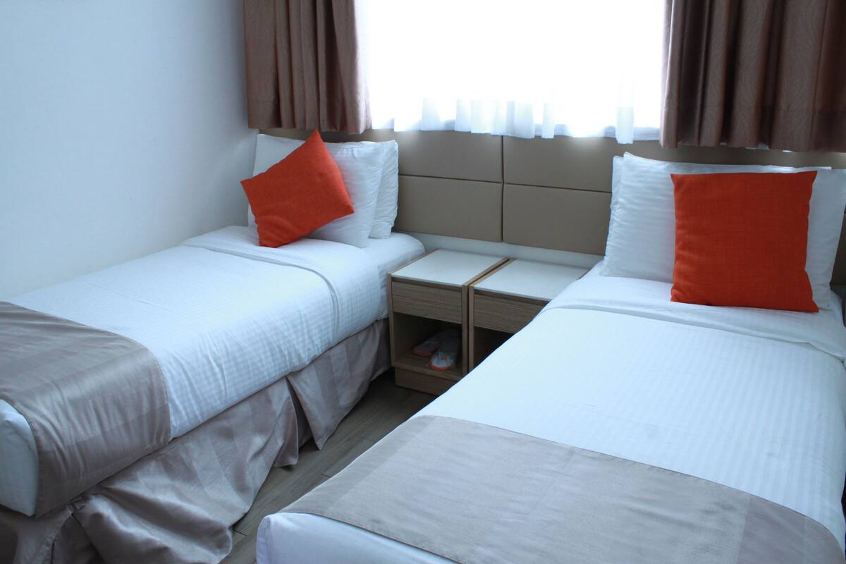 A hotel room with two beds, each with white bedding and orange pillows, a small bedside table, and a nightstand with a shoe rack.