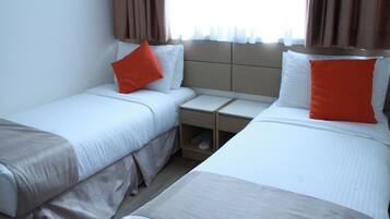 A hotel room with two beds, each with white bedding and orange pillows, a small bedside table, and a nightstand with a shoe rack.