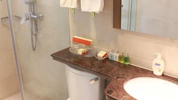 A bathroom with a toilet, sink, and shower. There are towels stacked on a shelf, soap dispensers, and a trash can.