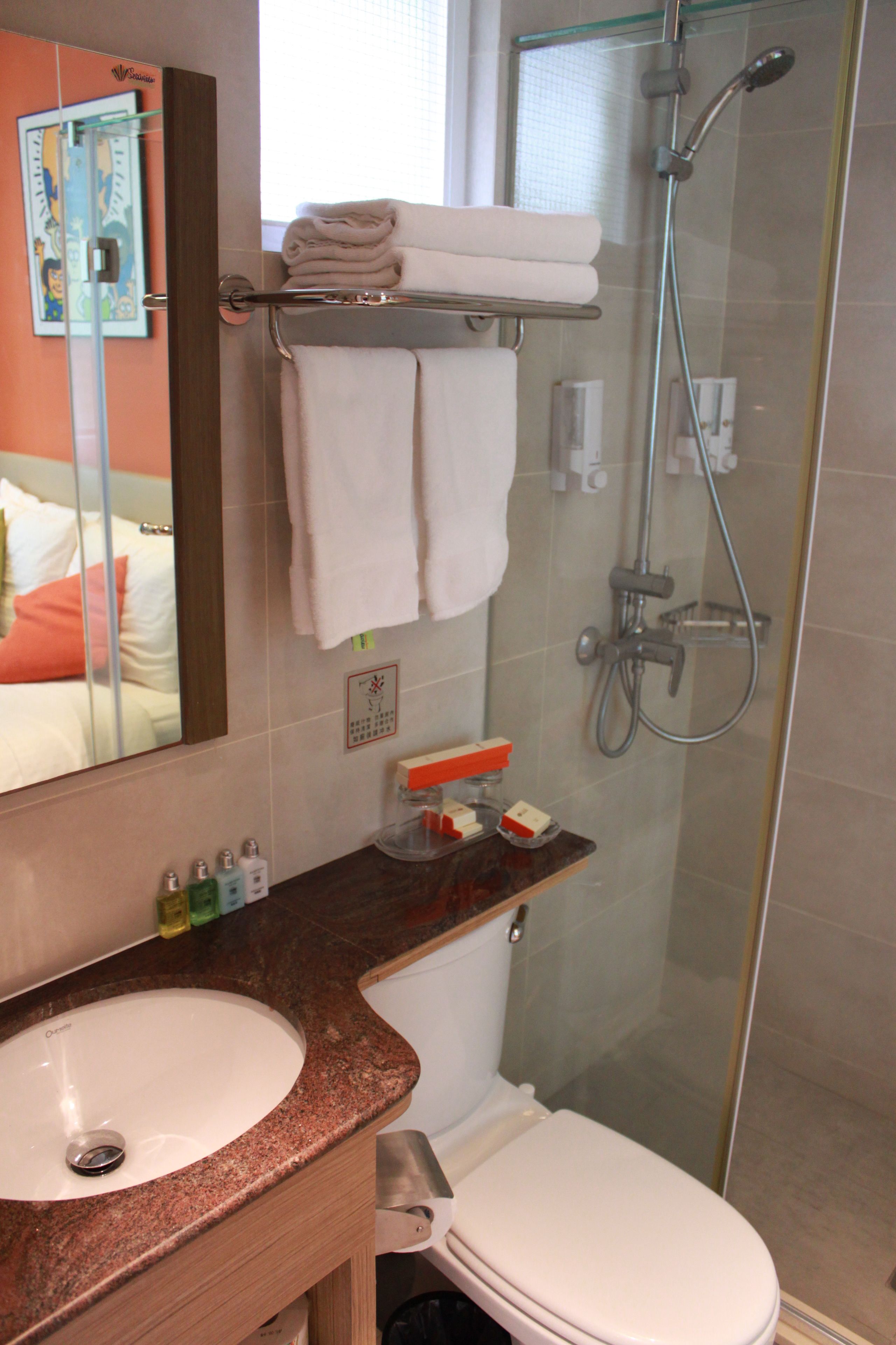 A bathroom with a sink, toilet, and shower. There is a towel rack with folded towels, a mirror, and toiletries on the counter.