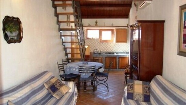 A cozy living area with a striped sofa, a dining table with chairs, a staircase, and a kitchen in the background. - Casa Ifigenia (Cefalù)