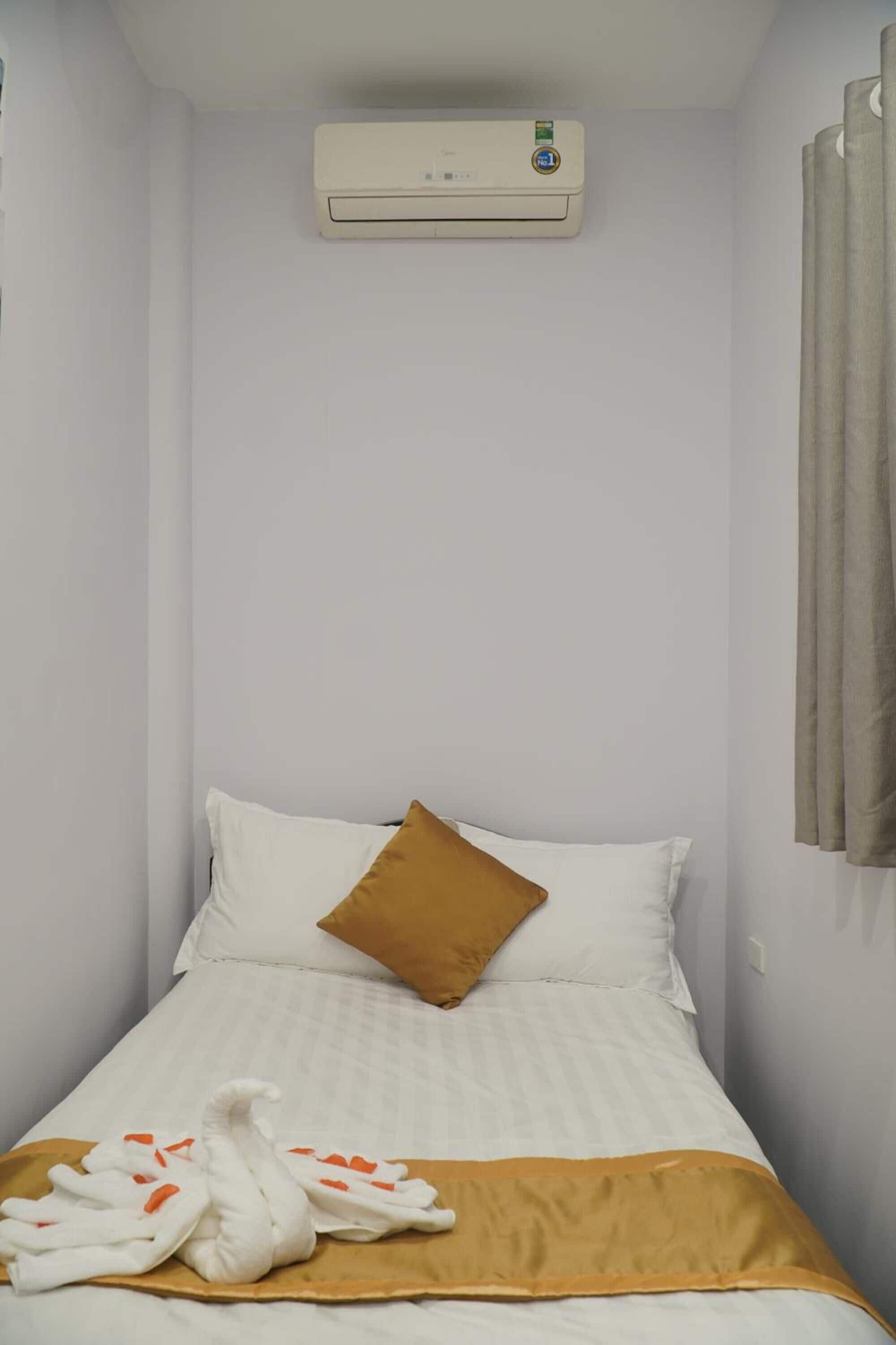 A small, single-bed room with a white bedspread, a brown pillow, and a towel shaped like a swan on the bed.