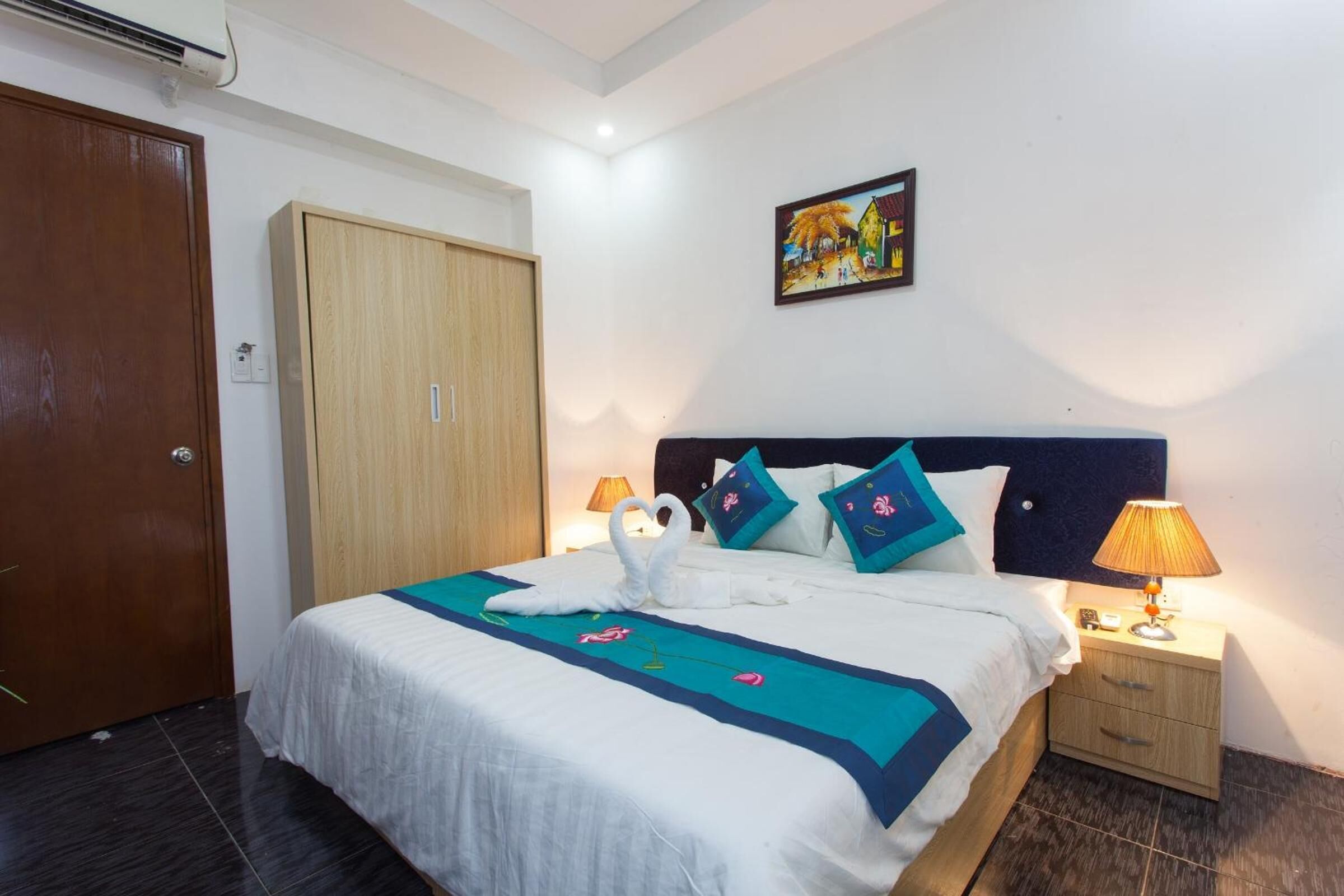 A neatly made bed with a blue and white bedspread, a wooden wardrobe, a nightstand with a lamp, and a painting on the wall.