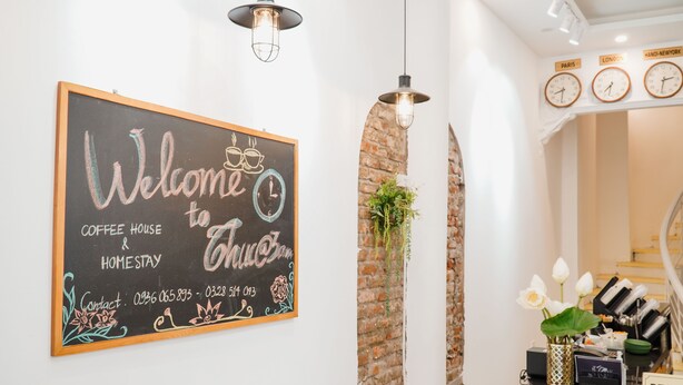 A reception area with a chalkboard sign reading "Welcome to Coffee House & Home Stay," a reception desk with a vase of flowers, and clocks on the wall indicating different time zones.