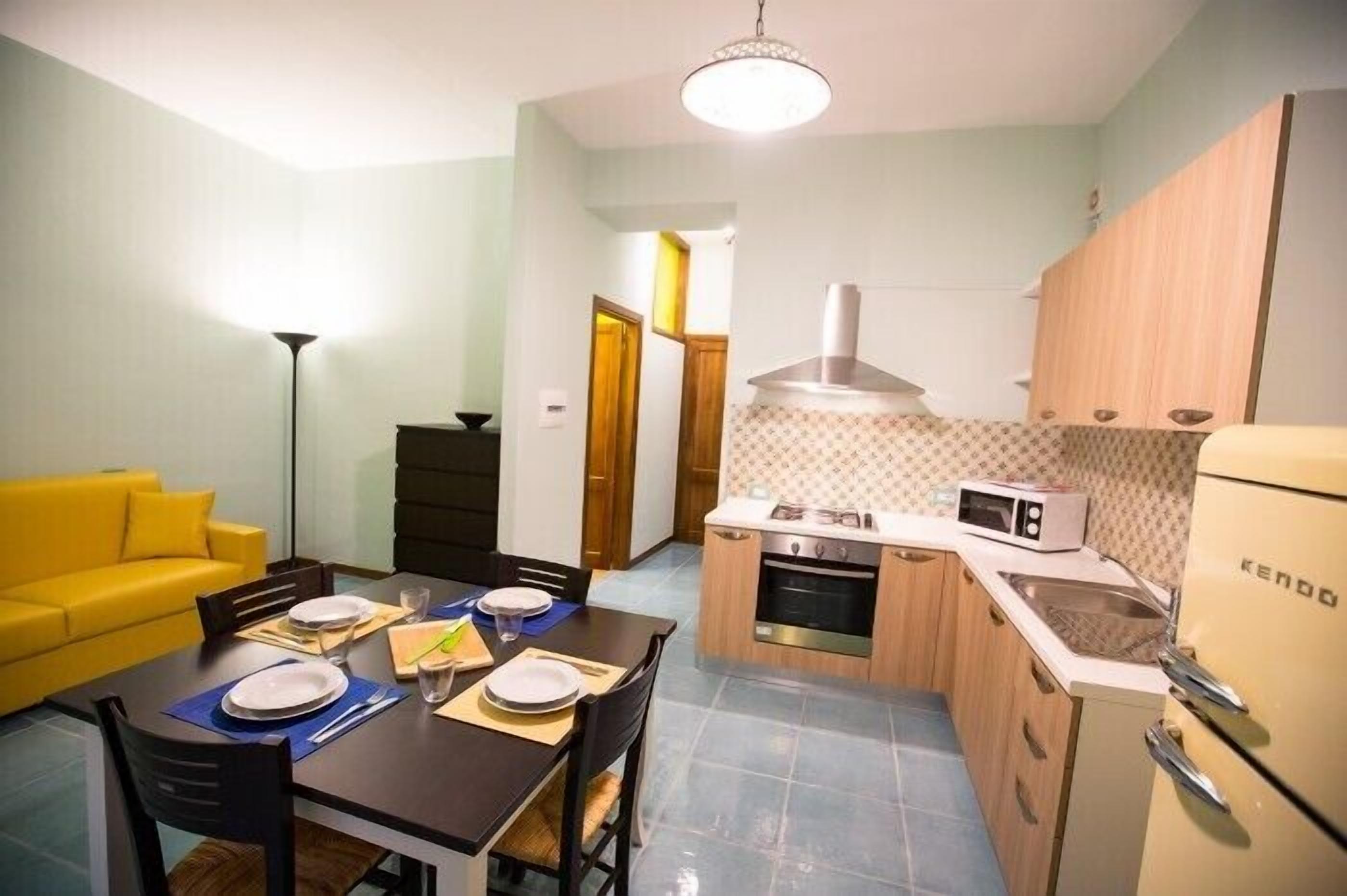 A compact kitchen with a stove, microwave, and a refrigerator. There is a dining table with chairs and a yellow sofa in the living area.