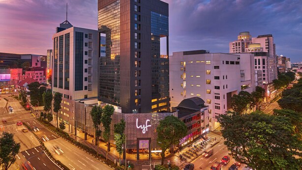 Un hotel moderno con un llamativo letrero 'lyf', rodeado de edificios urbanos y una calle concurrida al anochecer.