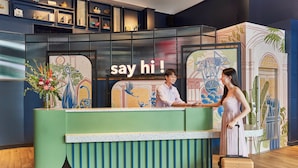 Una mesa de recepción con un cartel que dice "say hi!", un hombre y una mujer de pie frente al mostrador, y un suelo de madera.