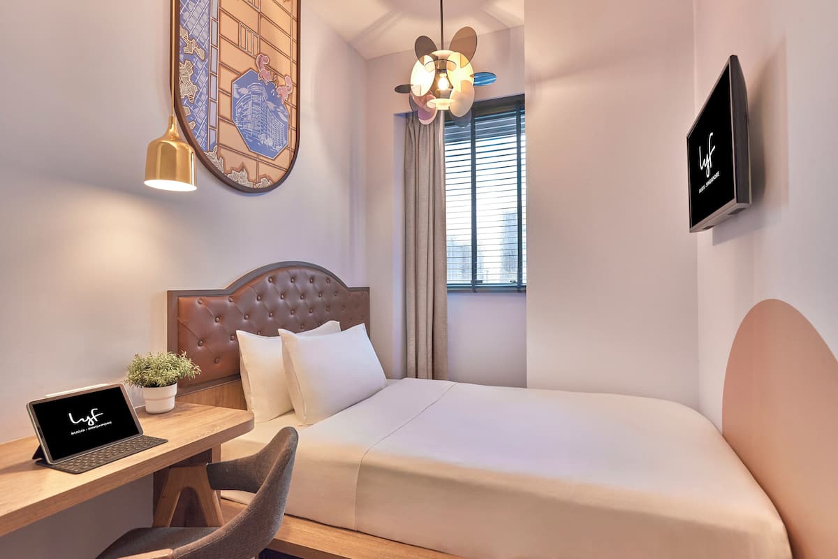 Une chambre d’hôtel avec un lit, un bureau avec un ordinateur portable, une plante en pot, un téléviseur fixé au mur et un ventilateur de plafond.