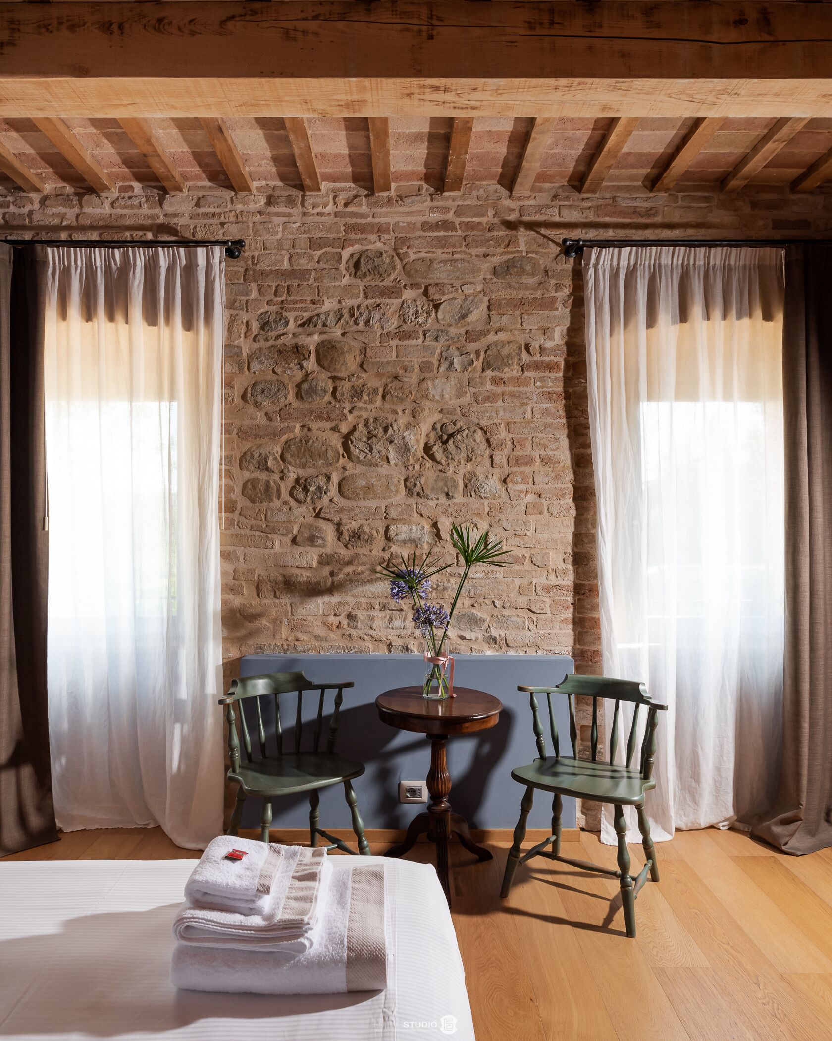 Una habitación rústica con paredes de ladrillo visto, vigas de madera, una mesa redonda de madera con dos sillas, un jarrón con flores y una pila de toallas en el suelo.