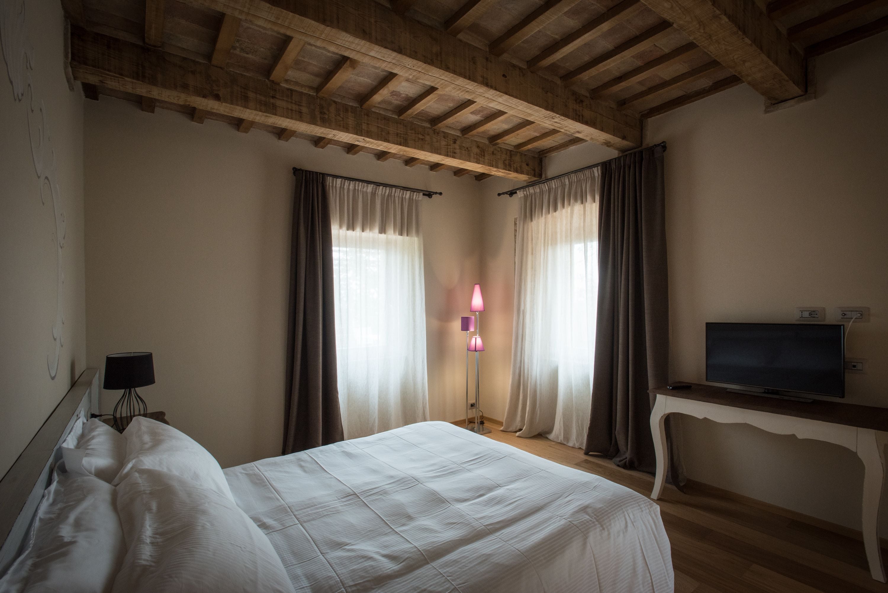 Une chambre avec un plafond en bois, un lit recouvert de draps blancs, une table de chevet avec une lampe et un téléviseur posé sur un meuble.