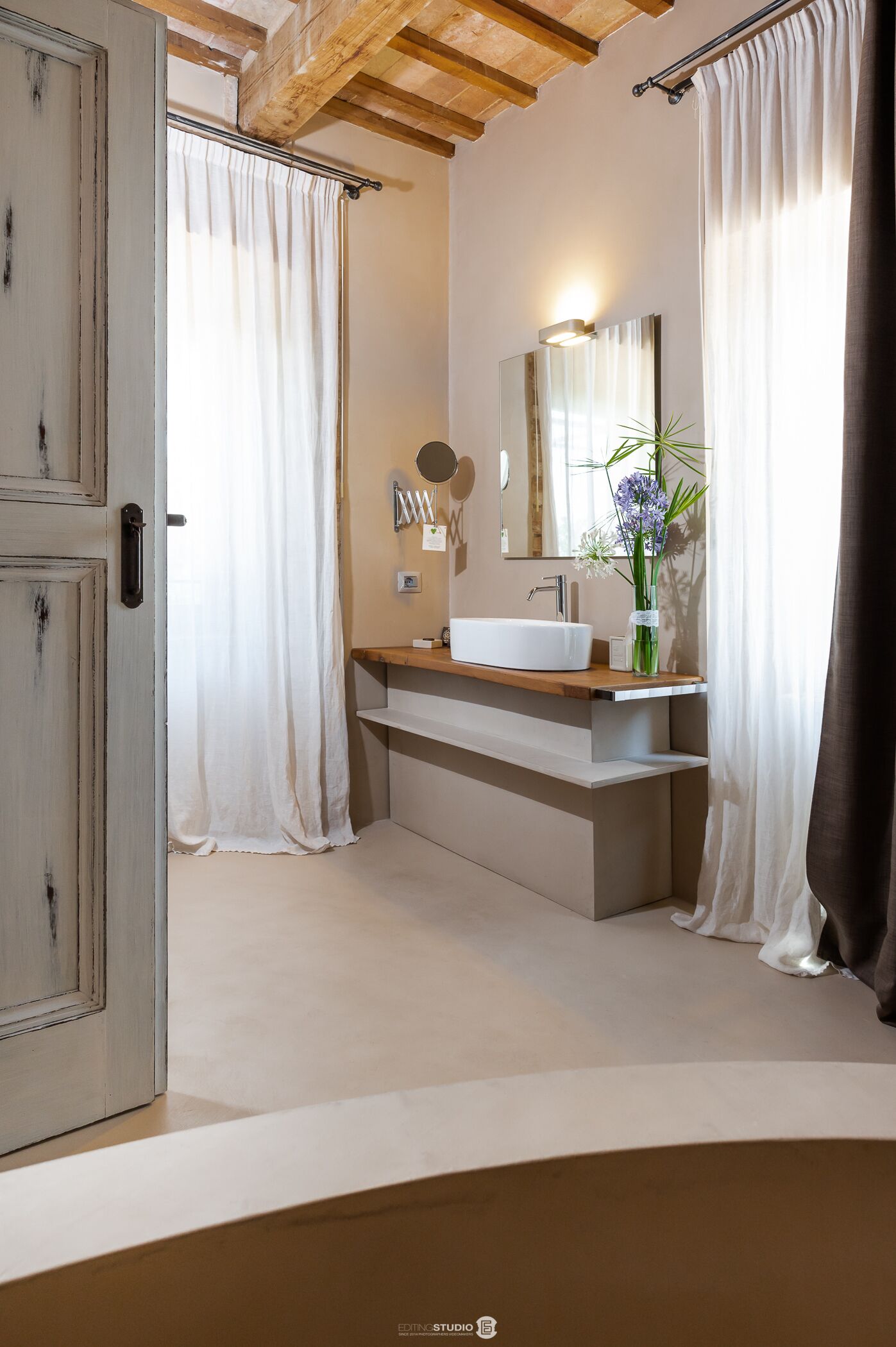 Un baño moderno con techo de madera, un espejo grande, un lavamanos blanco y un florero con flores.