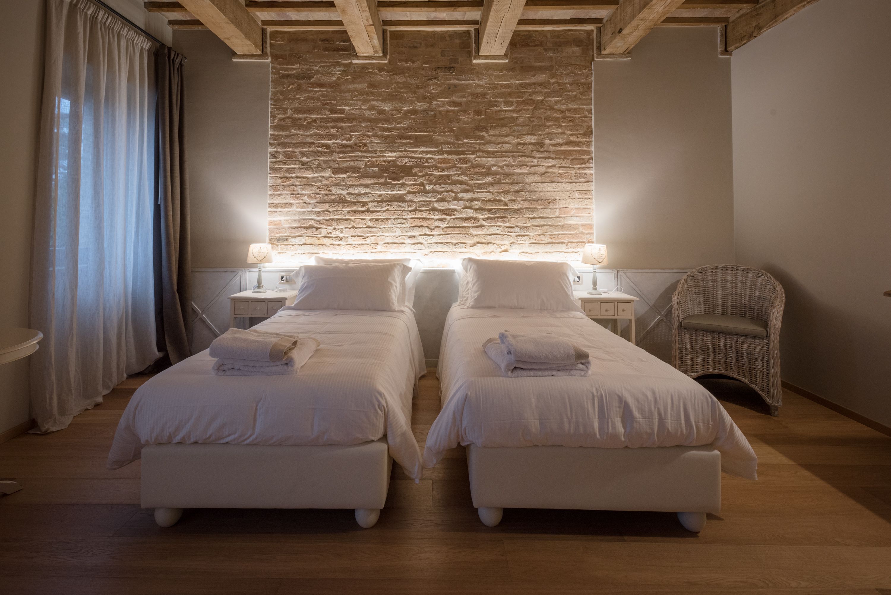 Un dormitorio con dos camas, una pared de piedra, vigas de madera en el techo y una silla de mimbre.