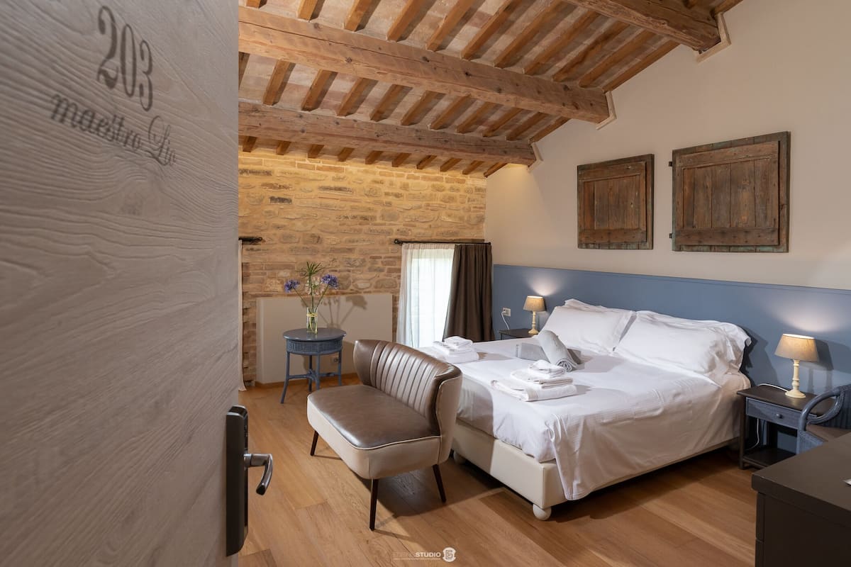 Un dormitorio con cama, una silla, una mesita de noche, una lámpara, una pequeña mesa con un florero y una puerta de madera con un número.