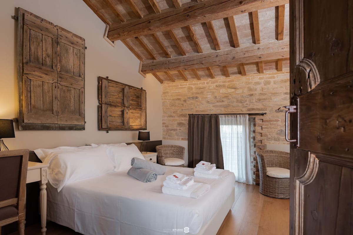 Un dormitorio con una cama grande, vigas de madera, paredes de piedra, sillas de mimbre y una puerta.