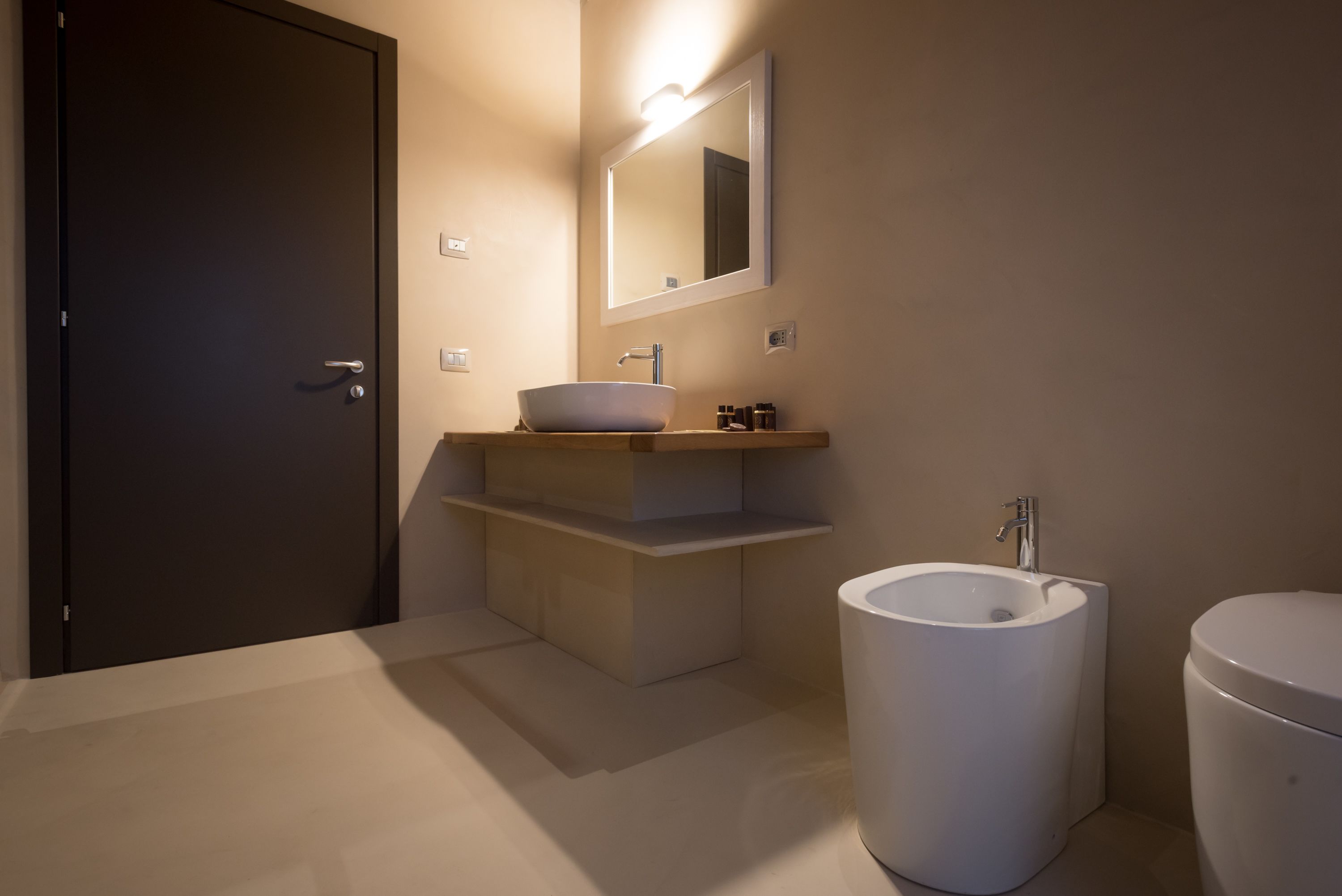 Un baño moderno con una puerta oscura, un lavabo blanco con lavamanos integrado y un inodoro independiente, además de un espejo con iluminación integrada.