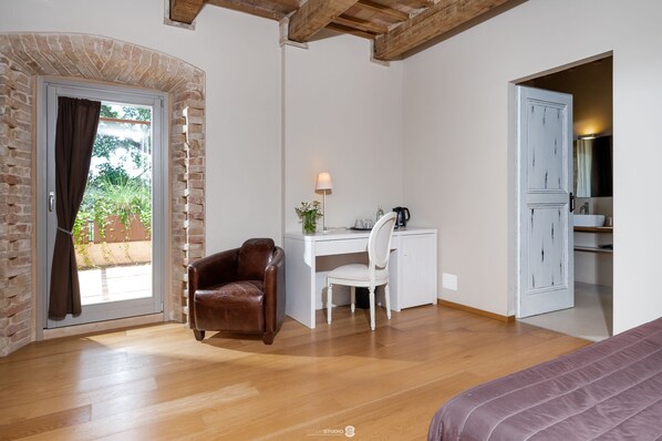 A room with a wooden floor, a desk with a chair, a leather chair, a bed, and a door leading to a bathroom. - Villa Anitori Prestige Relais & Spa (Loro Piceno)