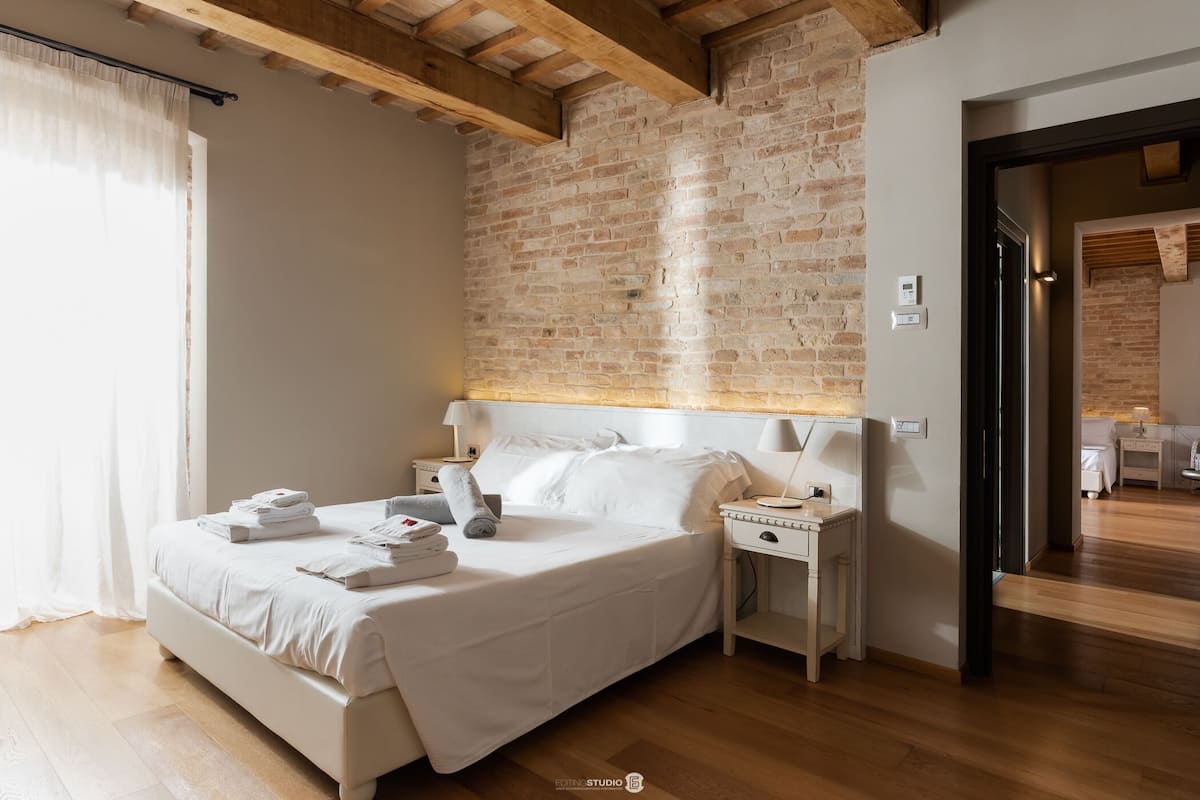 Un dormitorio con pared de ladrillo, vigas de madera, cama con ropa blanca, mesitas de noche con lámparas y suelo de madera.