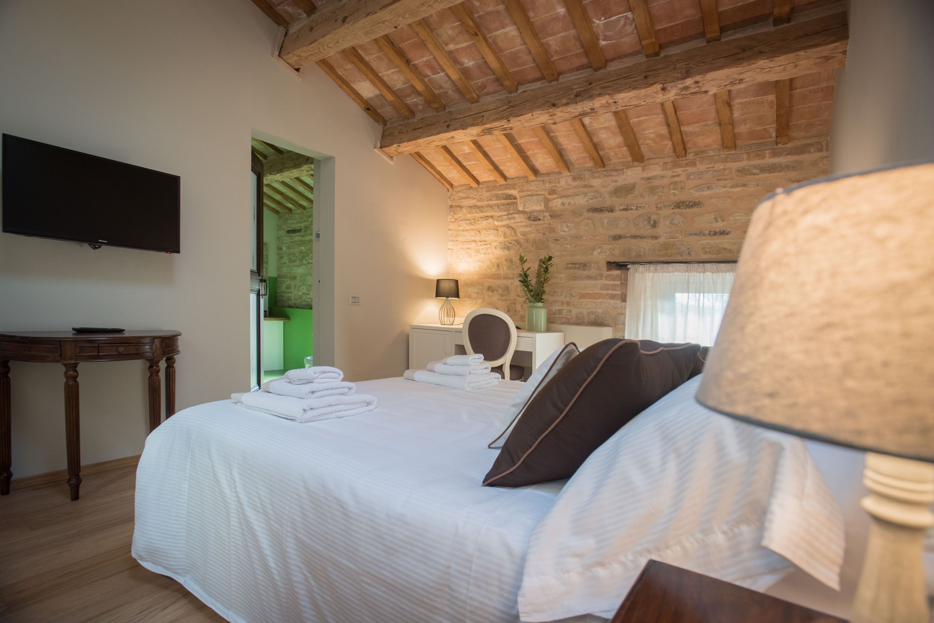 A bedroom with a large bed, a flat-screen TV mounted on the wall, a wooden table, and a brick wall.