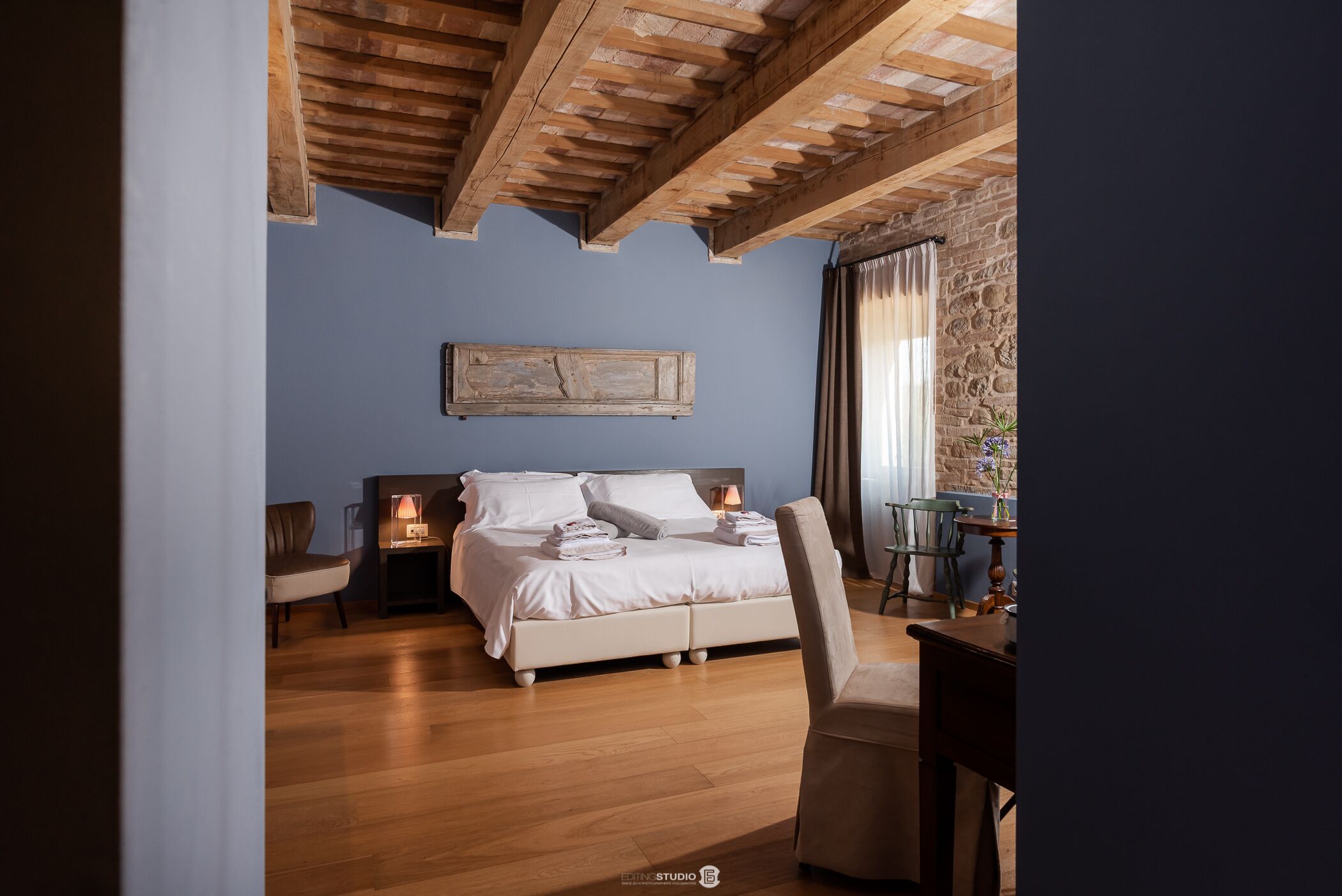 Une chambre avec un plafond en bois, un lit avec des oreillers et une lampe, une chaise et une table sur laquelle se trouve un vase.