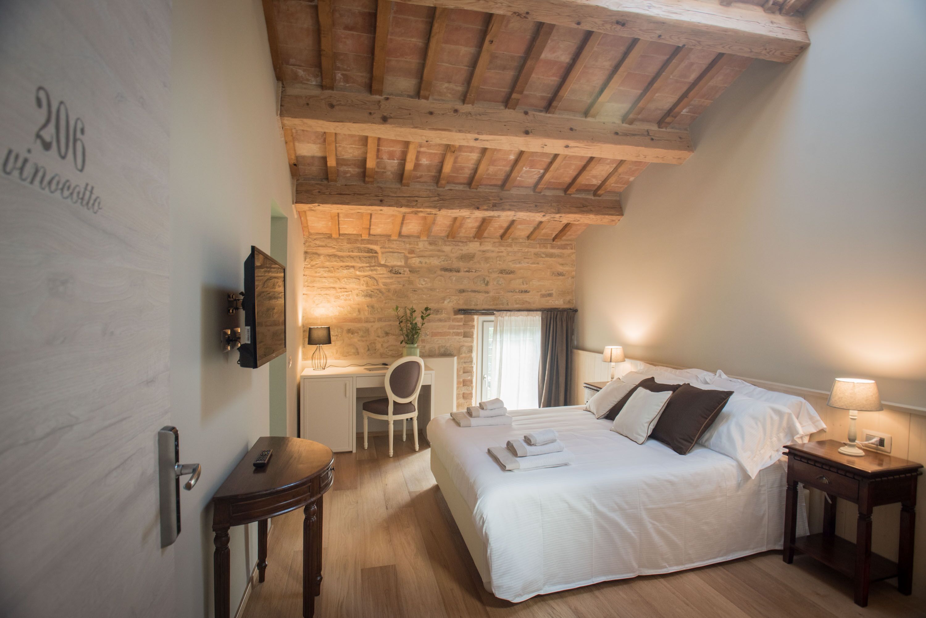 Ein Schlafzimmer mit Holzbalkendecke, einem Bett mit weißen Leinentüchern, einem Schreibtisch mit Stuhl und einem kleinen Tisch mit Lampe.