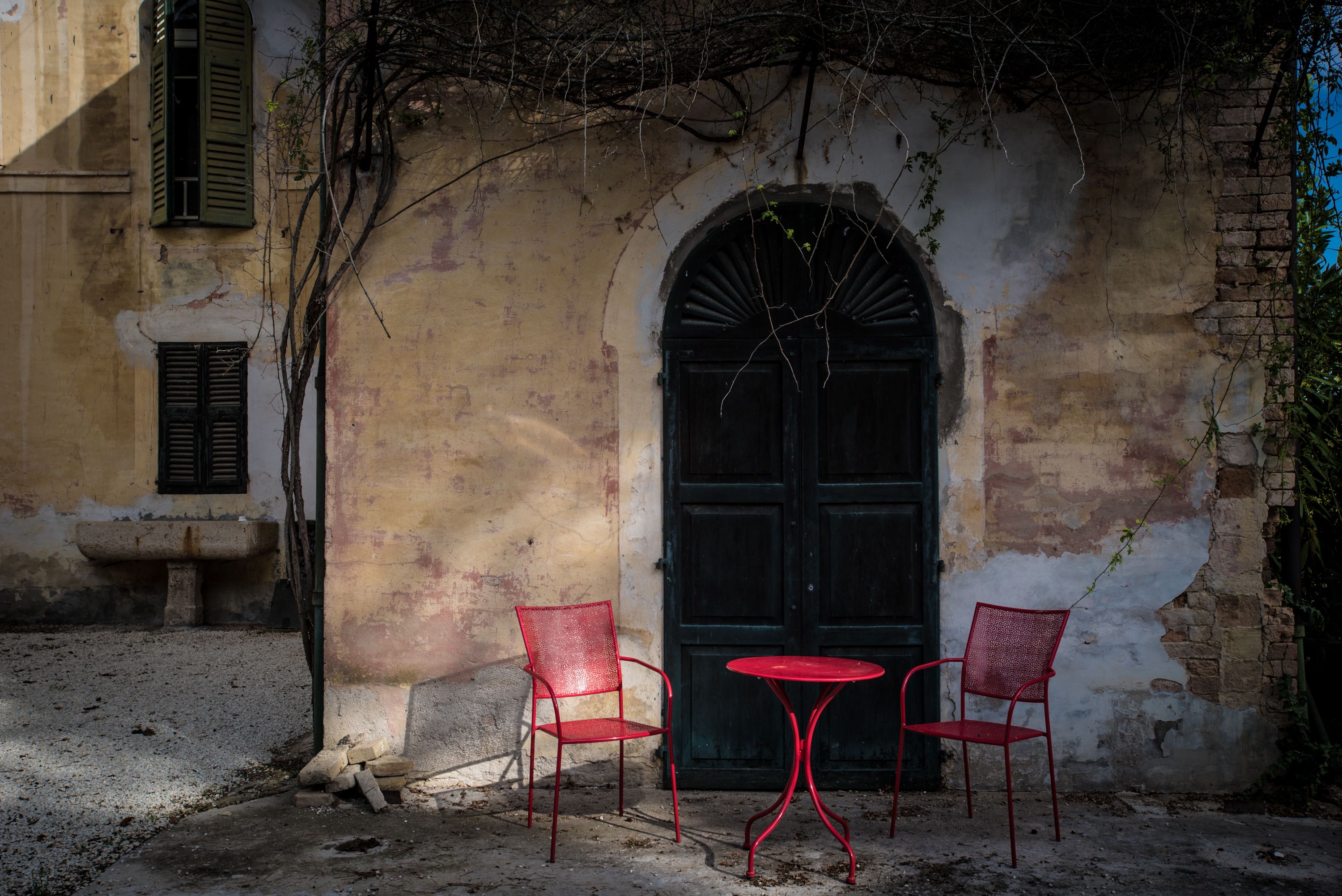 Un área de estar rústica al aire libre con una mesa redonda y dos sillas, un gran arco y un edificio con pintura descascarada y un banco de piedra.