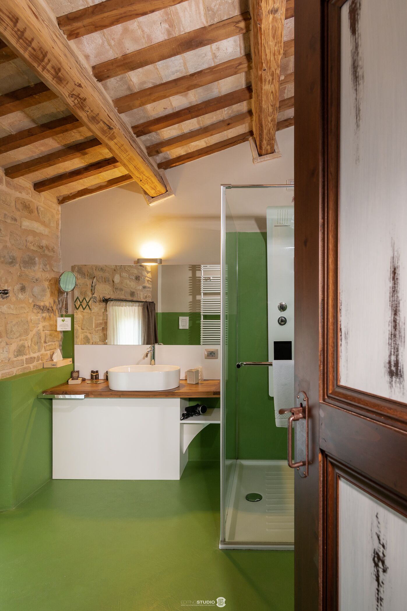 Une salle de bain avec un mur en pierre, des poutres en bois, un lavabo blanc, une douche verte et un miroir.