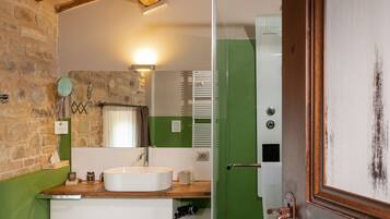 Une salle de bain avec un mur en pierre, des poutres en bois, un lavabo blanc, une douche verte et un miroir.