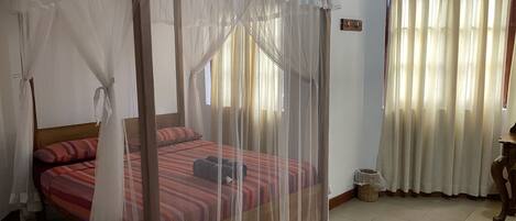 A room with a four-poster bed covered in a sheer canopy, a window with sheer curtains, and a tiled floor.