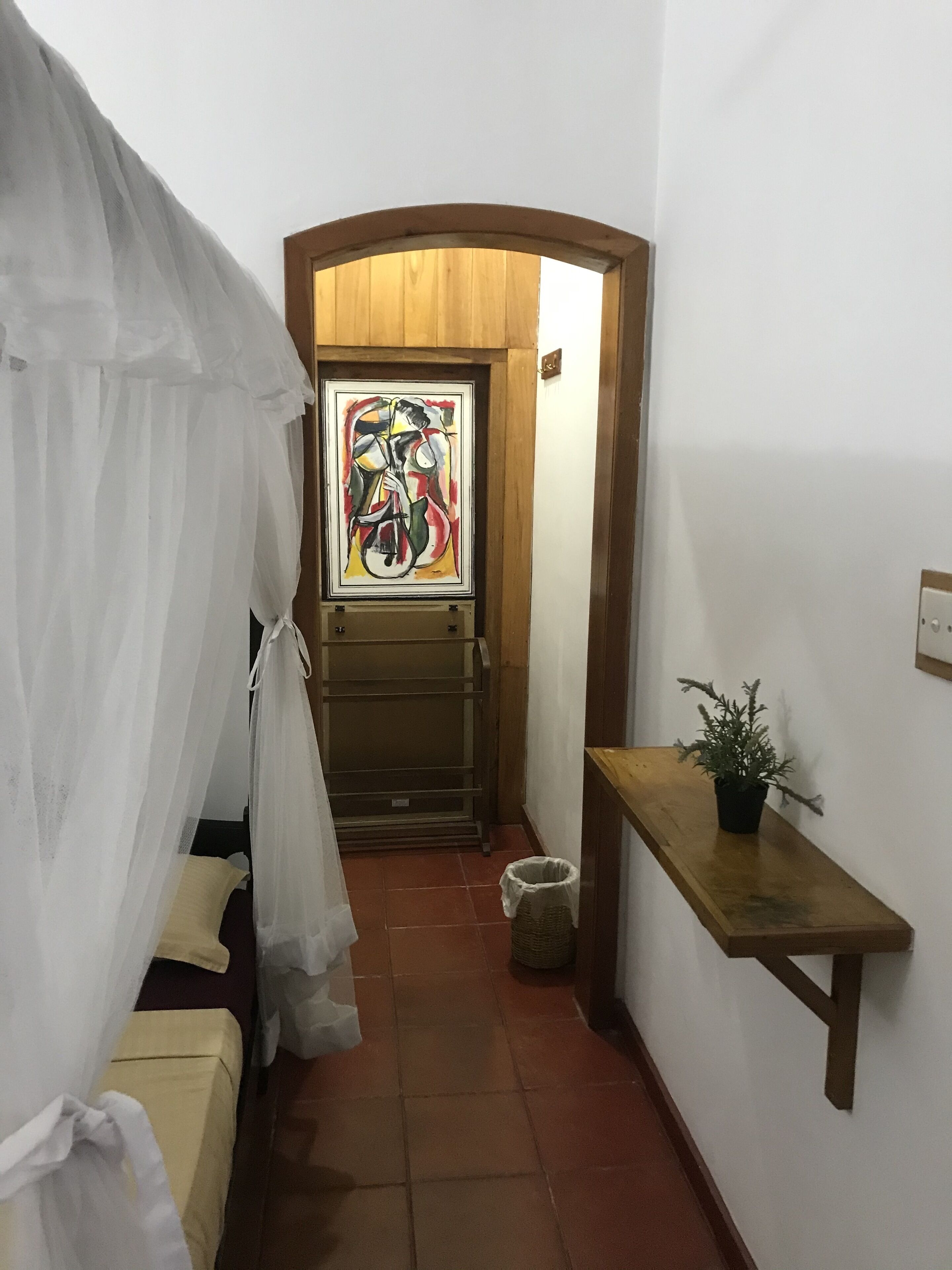 A narrow hallway with a bed on the left, a wooden bench on the right, and a door with a colorful painting above it.