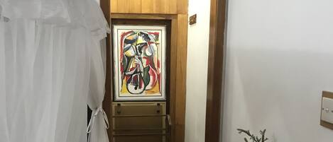 A narrow hallway with a bed on the left, a wooden bench on the right, and a door with a colorful painting above it.