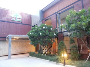 A building with a modern design, featuring a glass facade and wooden elements. There are two trees in the foreground, one with white flowers. - Buaksee Resort (Sing Buri)