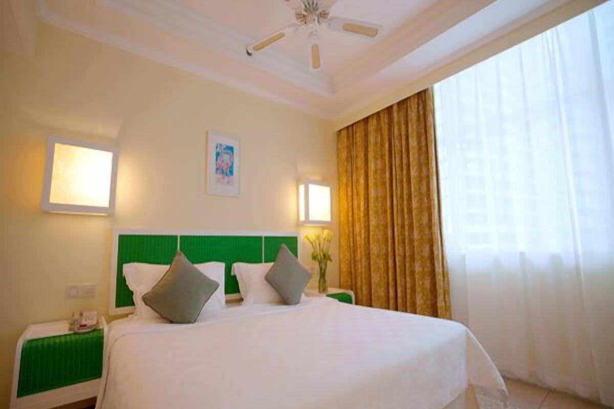 Habitación de hotel con una cama, cabecera verde, dos mesitas de noche, ventilador de techo y una ventana con cortinas.